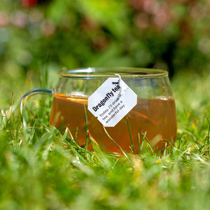 A glass cup of herbal tea sits on lush green grass, with a tea bag label reading, 'Dragonfly tea – Today, I'll drink tea and have a wonderful day.' The scene is sunlit, with a blurred natural background.