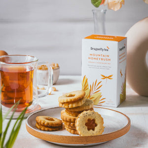 Sandwiched Mountain Honeybush Tea cookies with a star cut out showing amber-coloured apricot filling are stacked on a ceramic plate beside a box of Dragonfly Mountain Honeybush Tea and a glass mug of amber tea